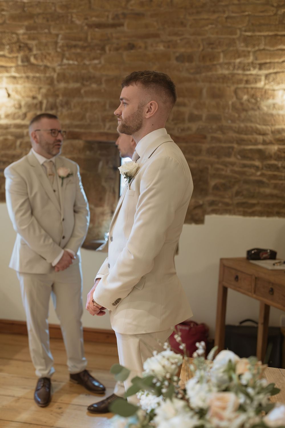 Two men wearing cream-colored suits stand in a rustic brick-walled room with floral arrangements on a wooden table.