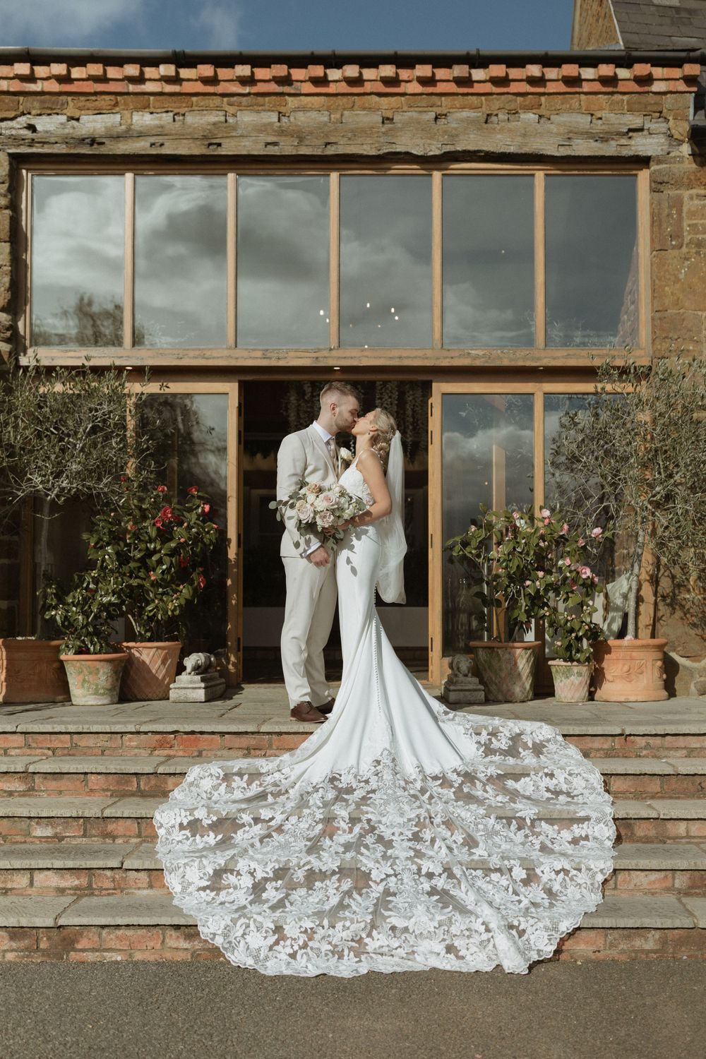 Newlyweds embrace on stone steps with a stunning long-train lace wedding gown against a rustic greenhouse backdrop.