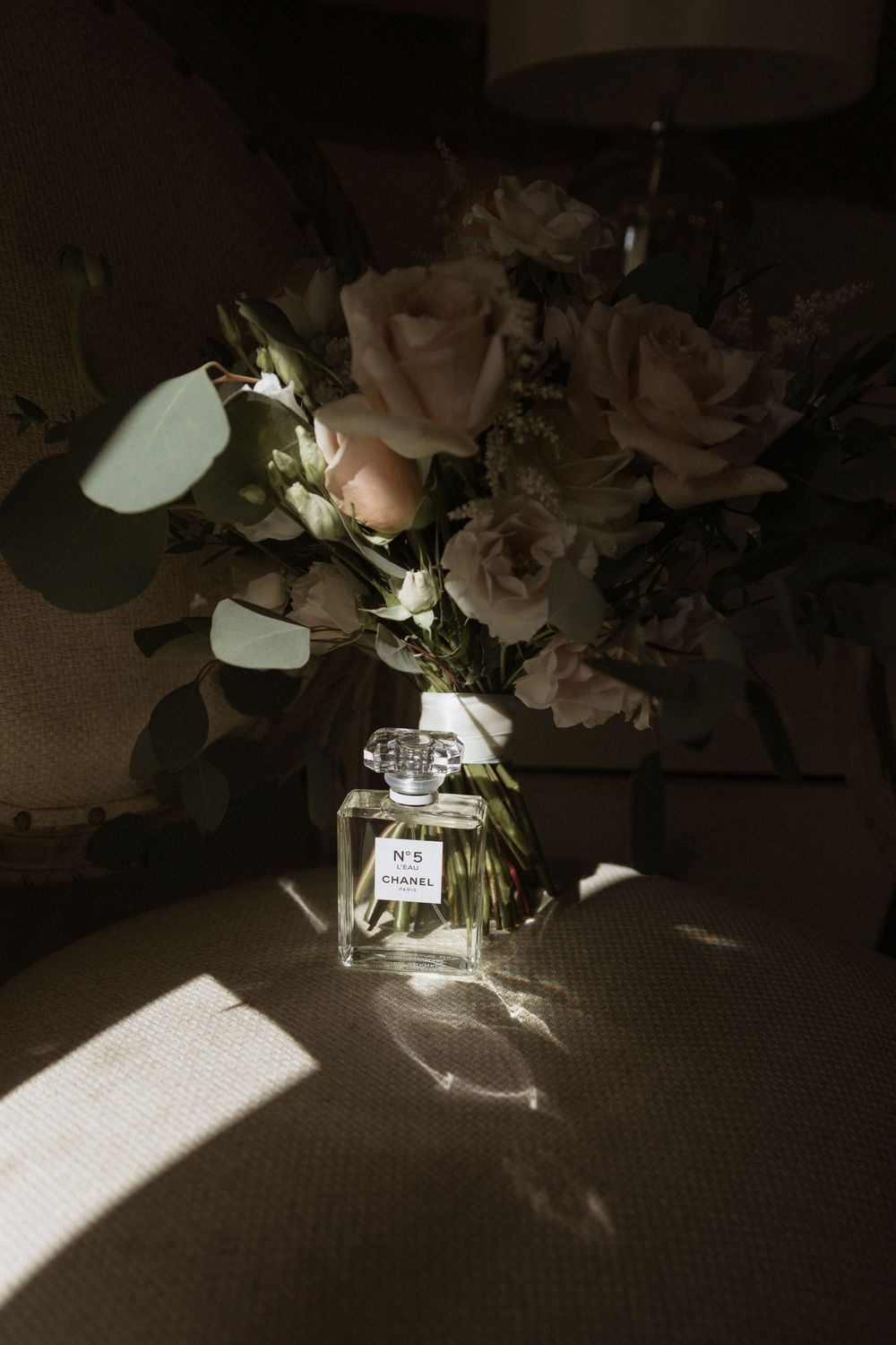 A Chanel No. 5 perfume bottle sits beside delicate pink roses in dramatic lighting with strong shadows.