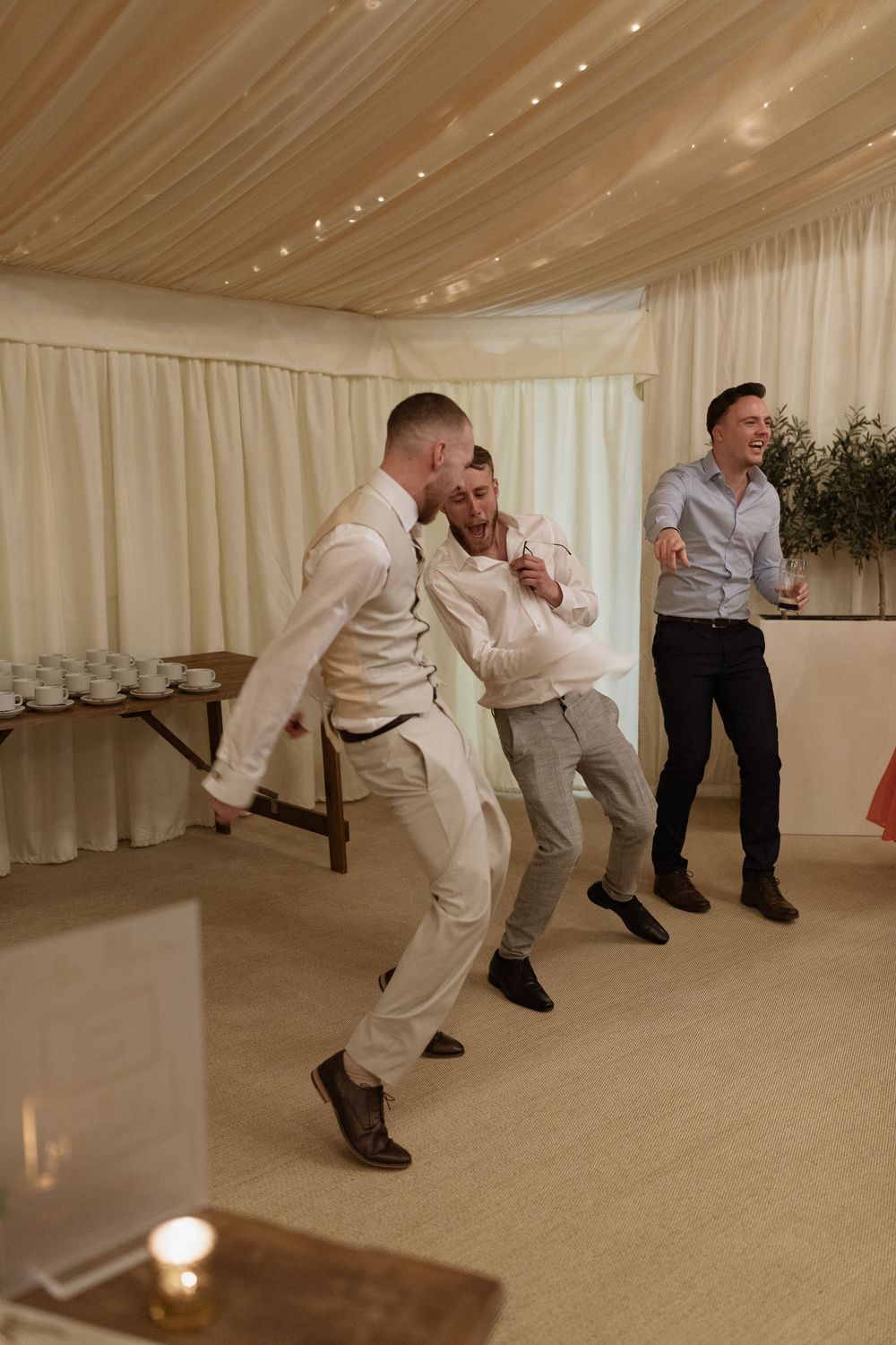 Three people dancing and laughing together in a reception tent with draped fabric ceiling and mood lighting.