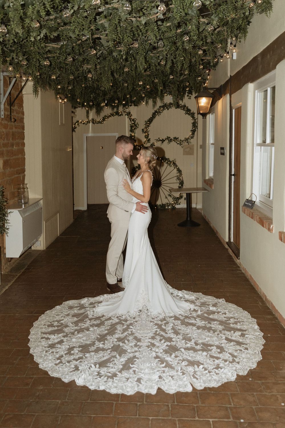 Elegant wedding couple poses in a rustic corridor adorned with overhead greenery and a flowing lace train.