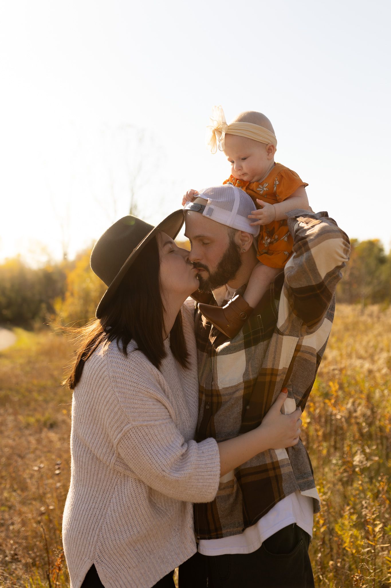 Family embraces in a golden autumn field during sunset while wearing cozy sweaters and hats.