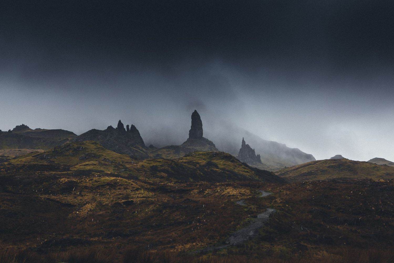 Dramatic rocky spires rise from misty Scottish highlands under moody storm clouds.