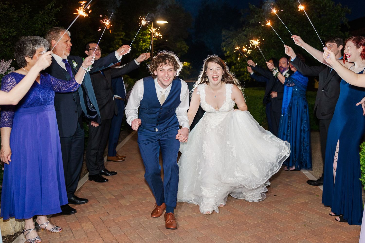 Wedding couple exits ceremony through sparkler tunnel lined with wedding party members in blue dresses and suits at night.