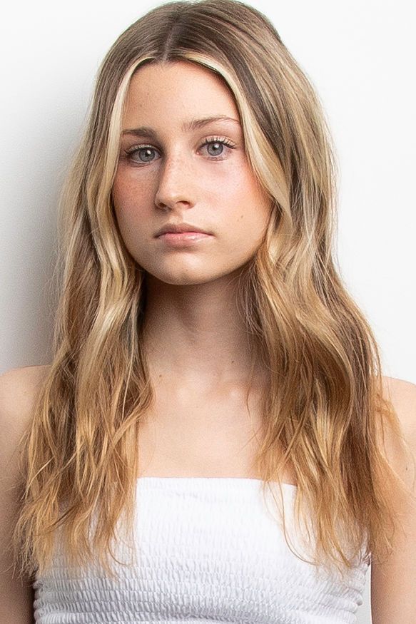Young woman in white top with long wavy honey blonde hair poses against white background in natural makeup look.