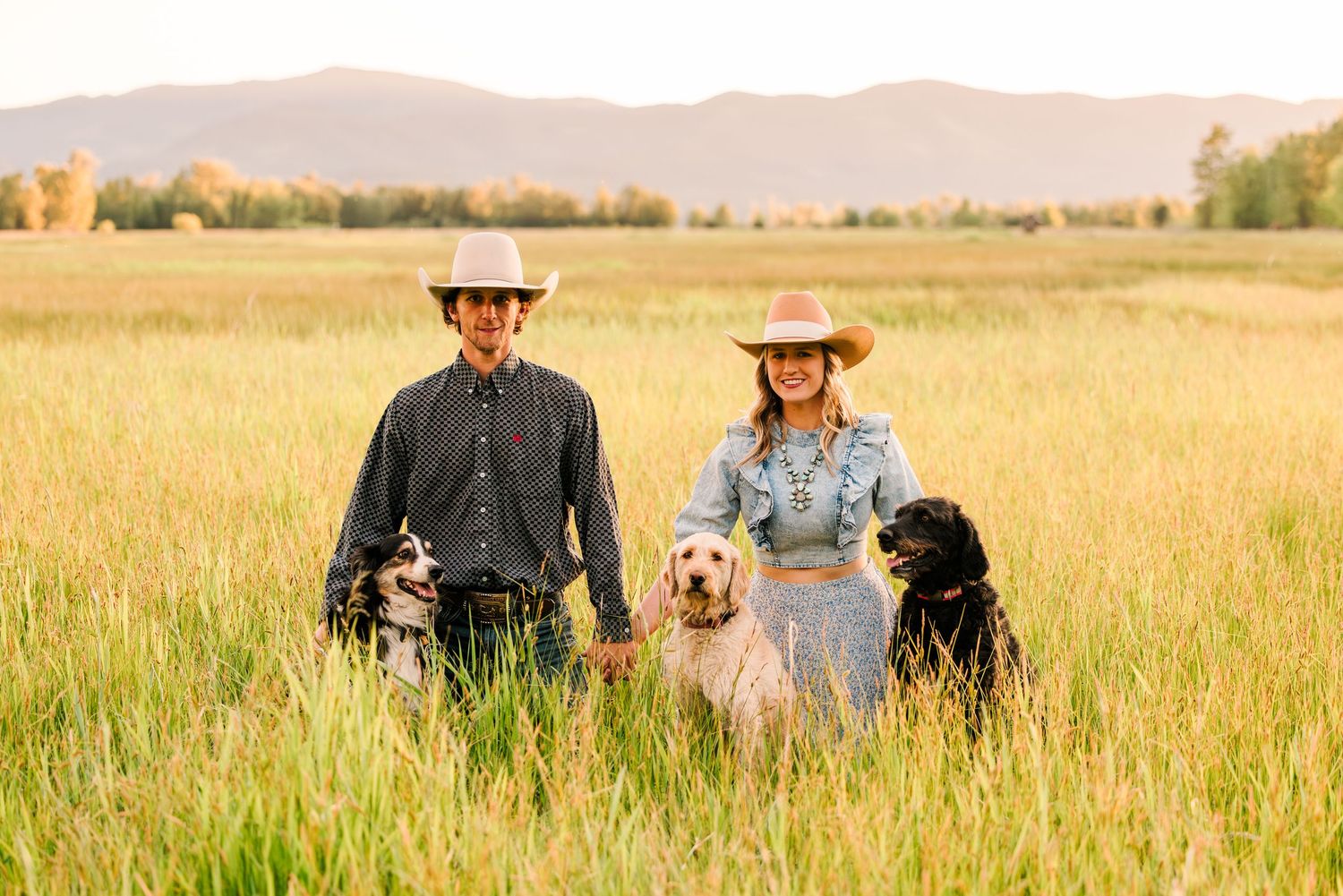 Two people in cowboy hats stand with their black and white dogs in a golden summer meadow with mountains in background.