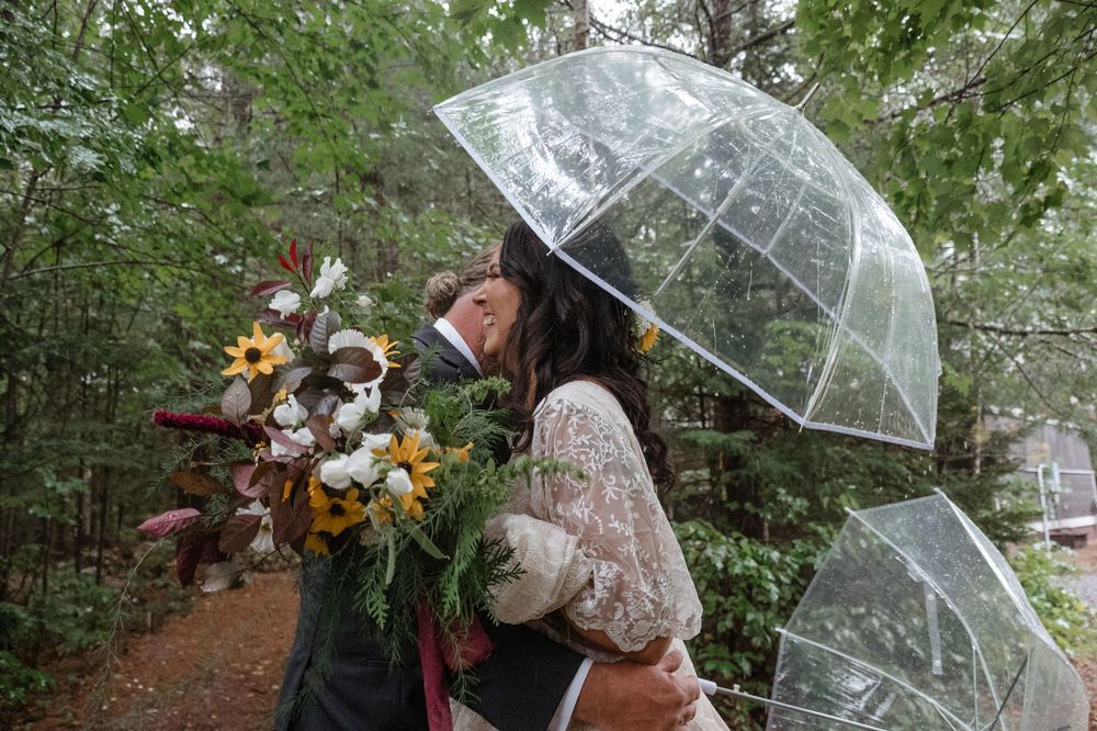 Two clear umbrellas protect people from rain as they embrace and hold a colorful autumn bouquet on a forest path.