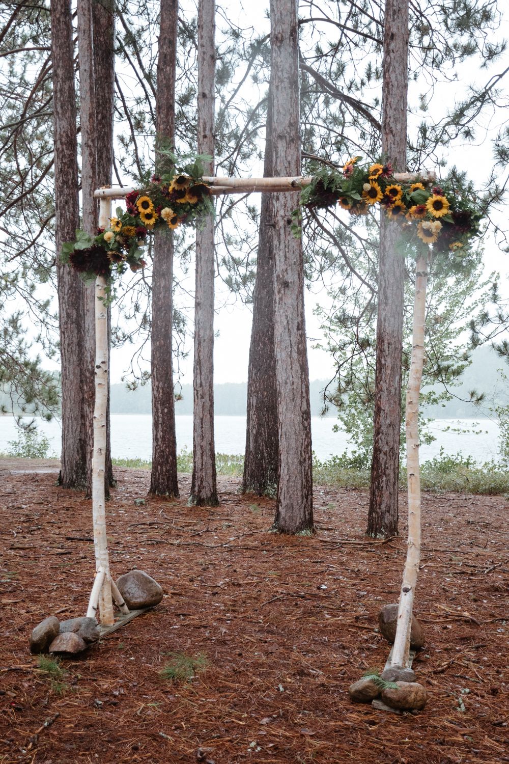 Pine forest wedding arch decorated with sunflowers and greenery against tall tree trunks.