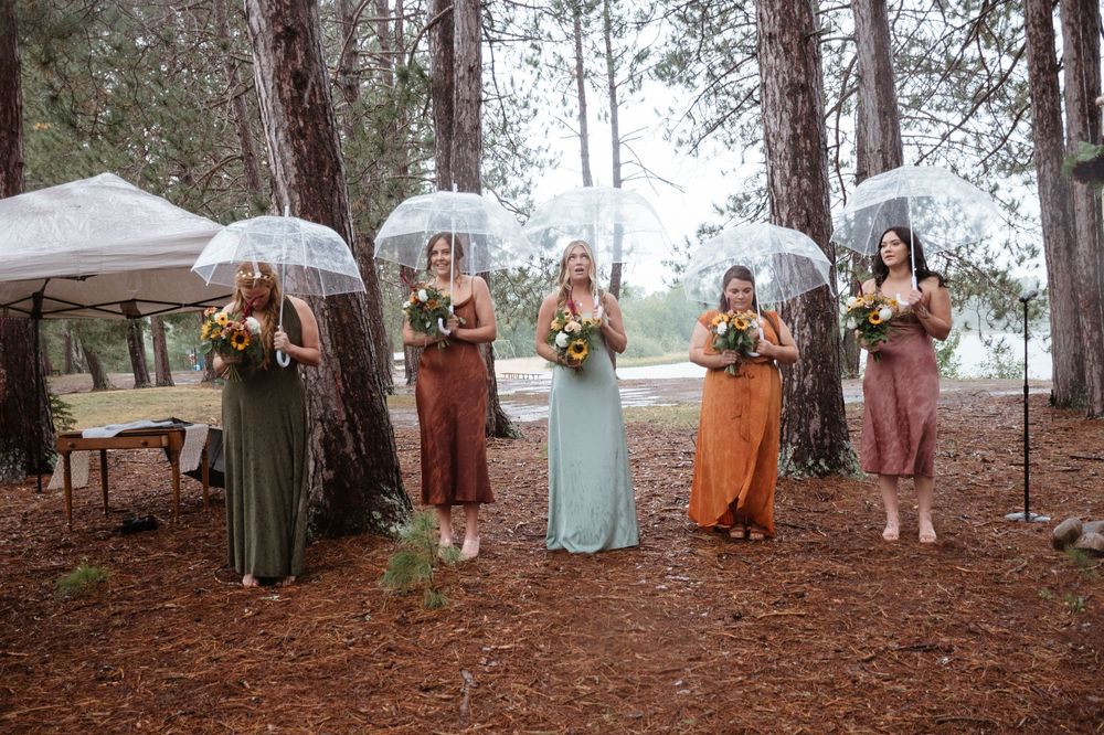 Bridal party holding clear umbrellas and bouquets stands in line on forest floor wearing autumn-colored dresses.