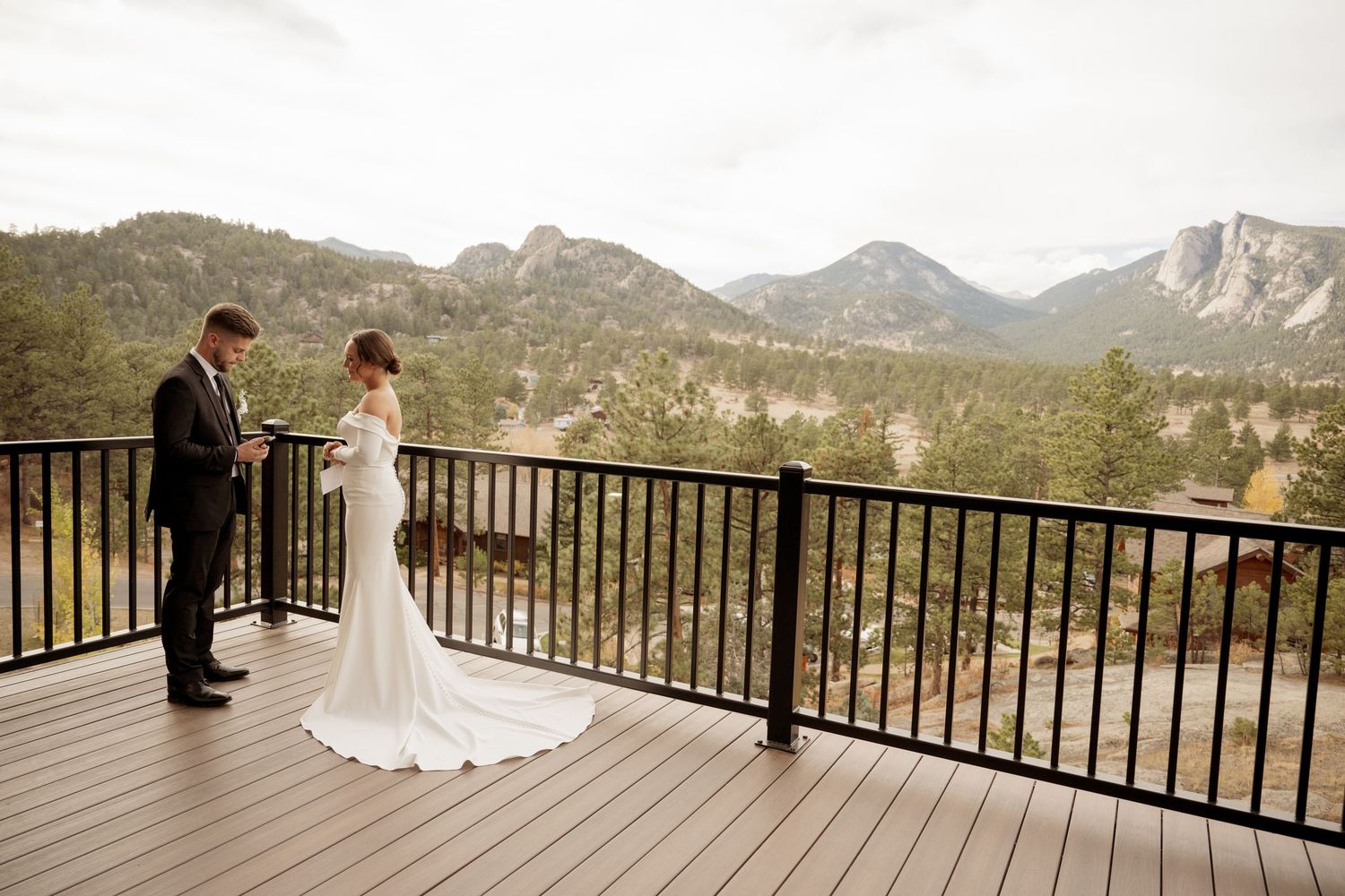 A couple walks hand in hand on a grassy hill overlooking majestic mountain ranges under cloudy skies.