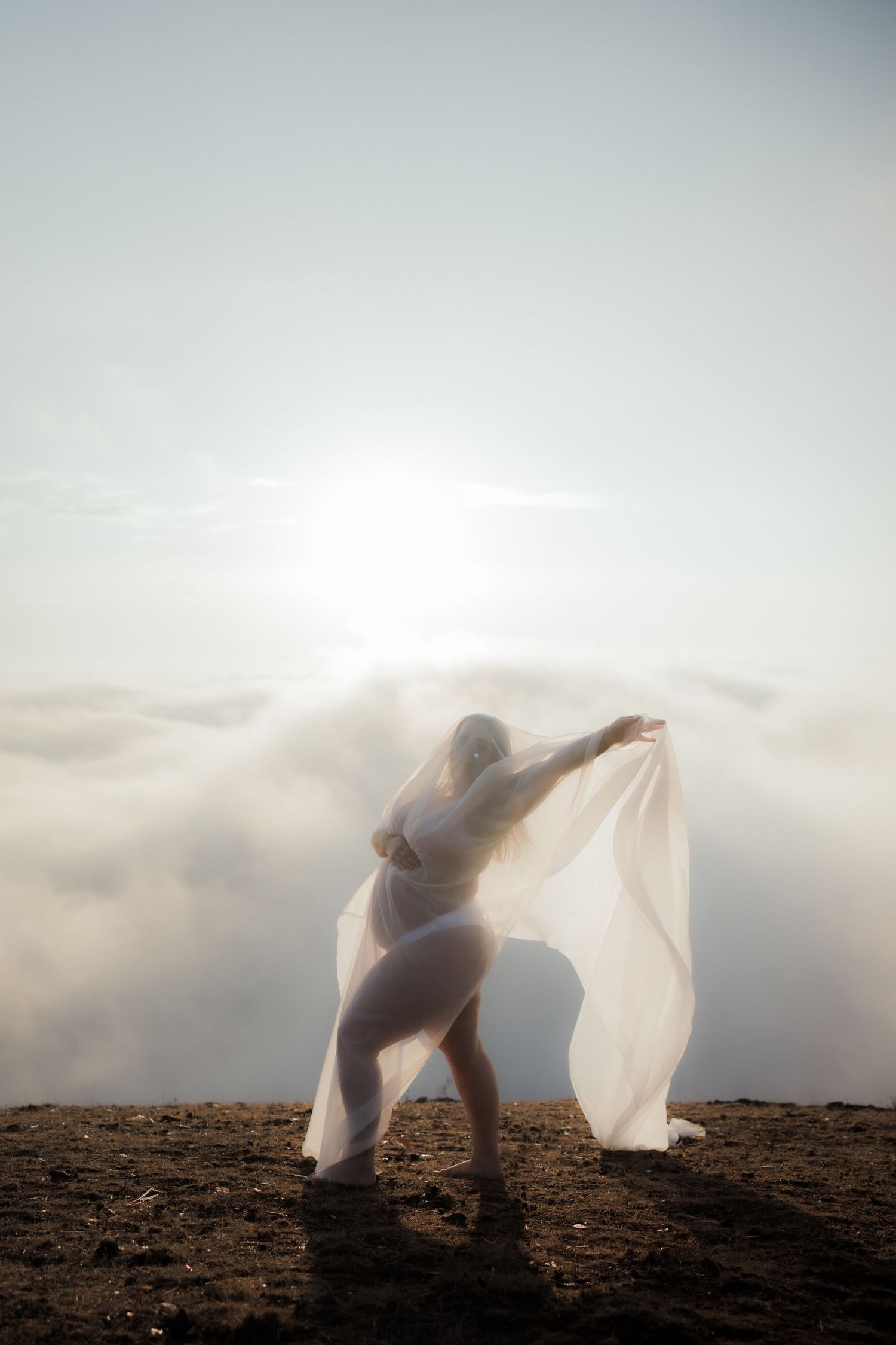 Ethereal silhouette dancing with flowing fabric against bright sunlit sky on mountain terrain.
