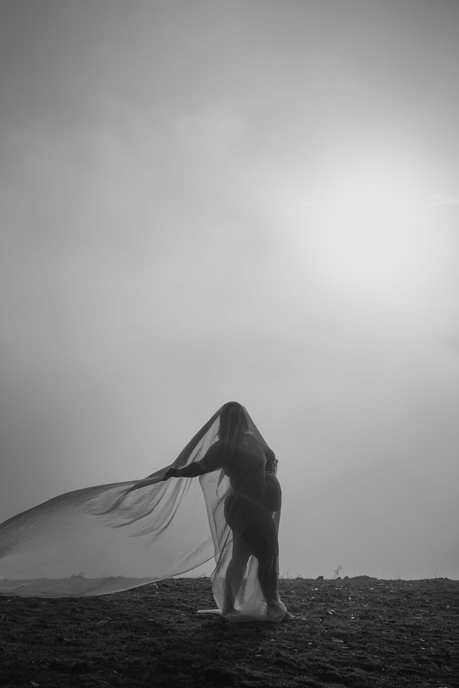 A silhouetted figure stands on a hilltop with flowing fabric billowing in the wind against a misty black and white sky.
