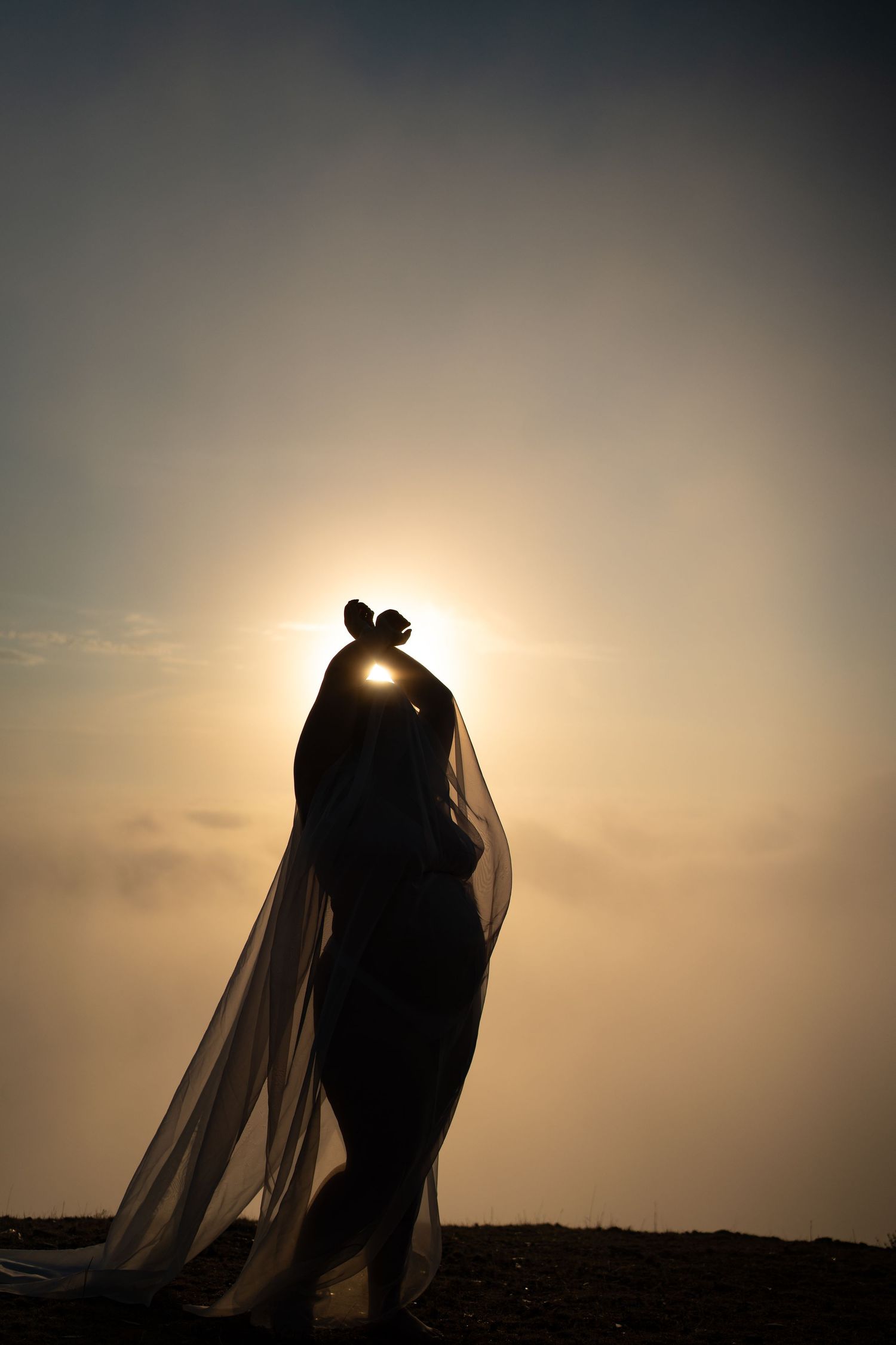 Silhouette of a graceful figure in flowing dress and veil against a golden sunset sky in dramatic backlight.