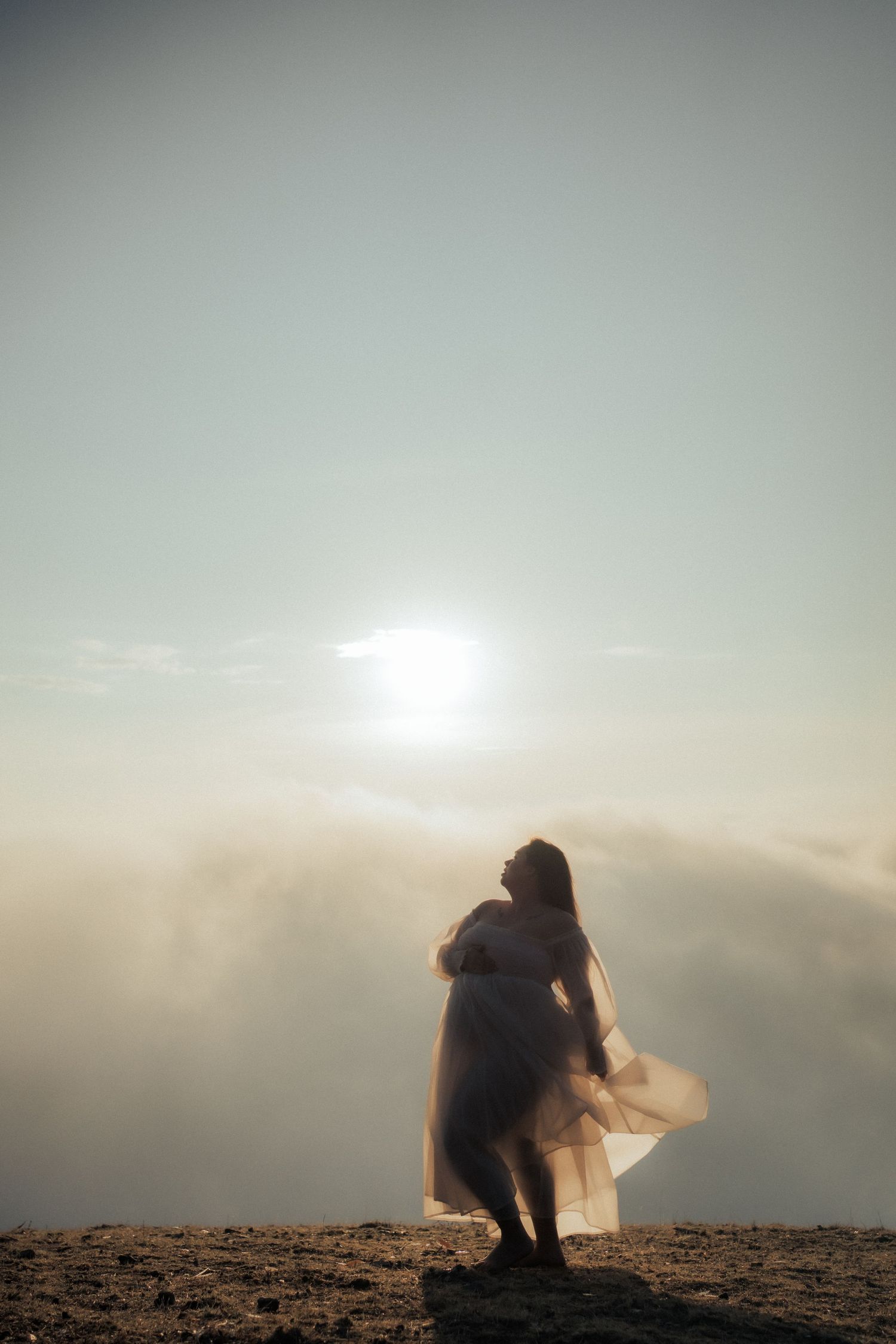 A silhouetted figure sits on a hilltop against a misty sunset sky as glowing sunlight pierces through the clouds.