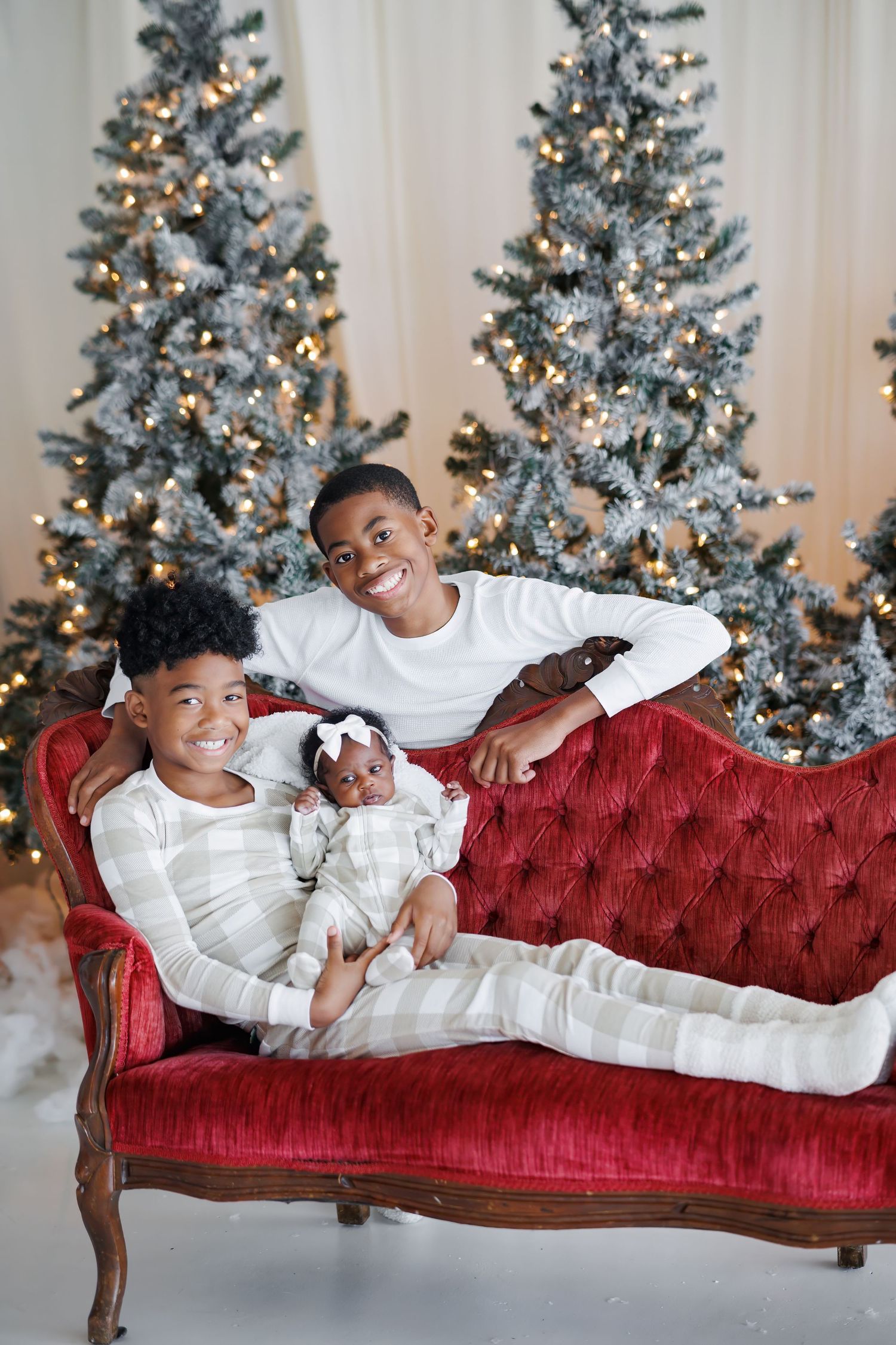 Two brothers in white pajamas lounging on a red vintage sofa with Christmas trees in the background during a Dallas holiday photoshoot