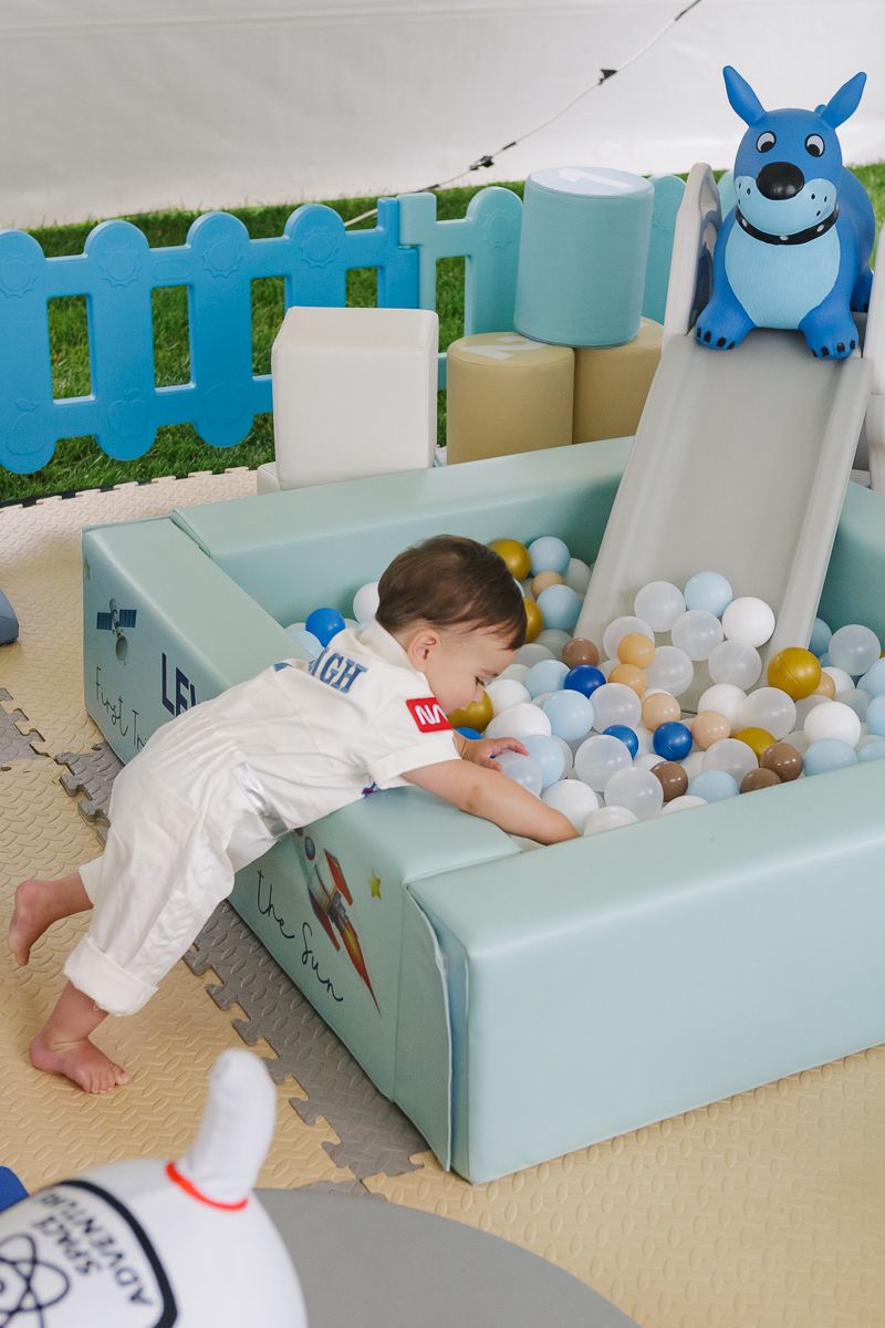 Adorable first birthday boy dressed as an astronaut, exploring the ball pit at his space-themed party in Manhasset.