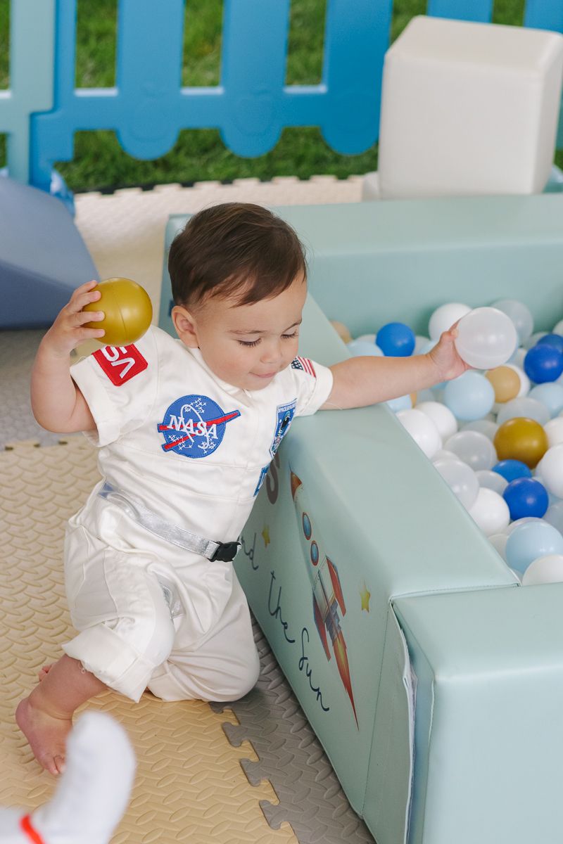 Joyful toddler in an astronaut suit laughing while playing in a ball pit, captured during his milestone birthday celebration