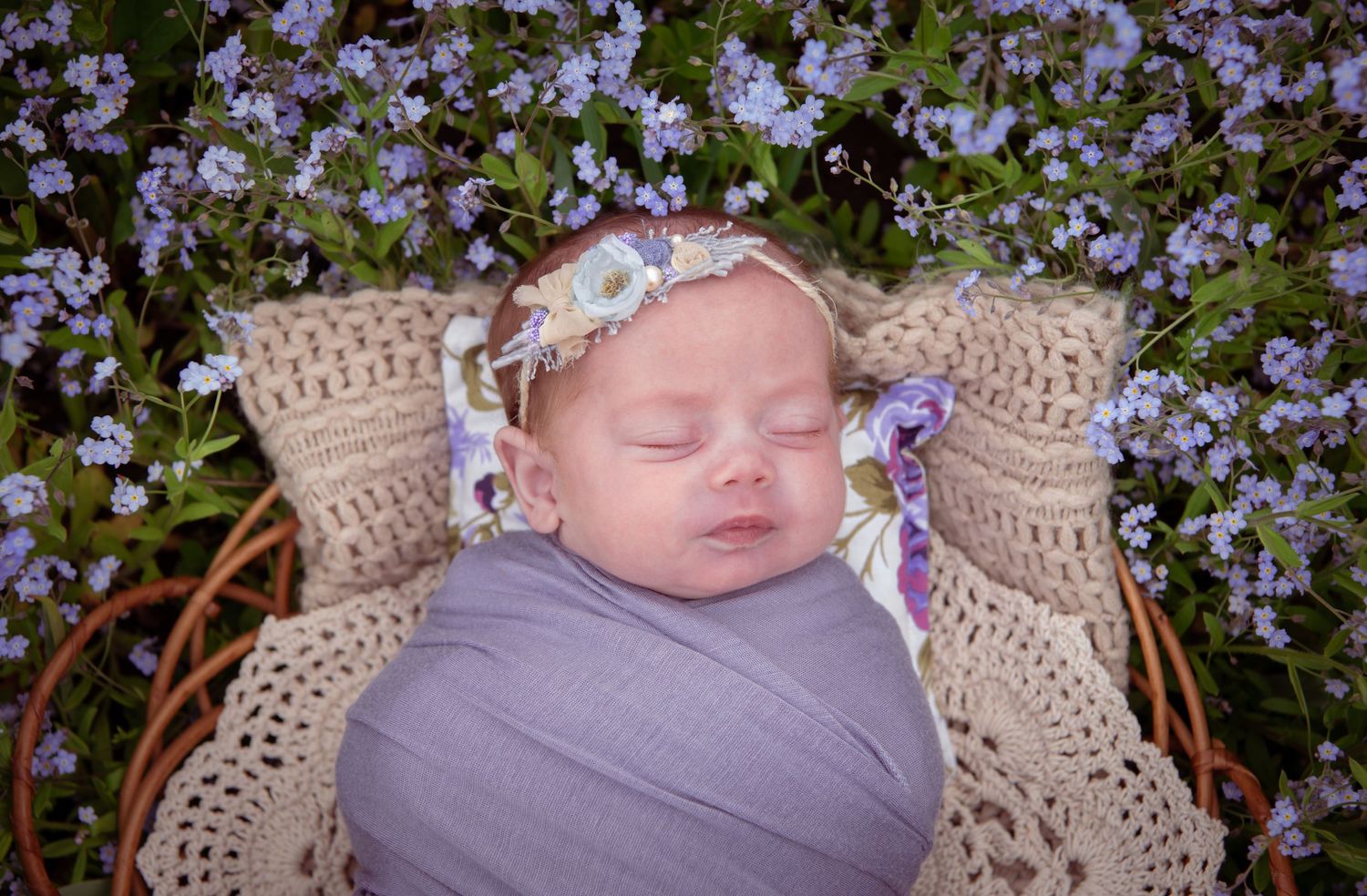 Sleeping baby wrapped in gray swaddle lying in wicker basket surrounded by purple flowers wearing floral crown.