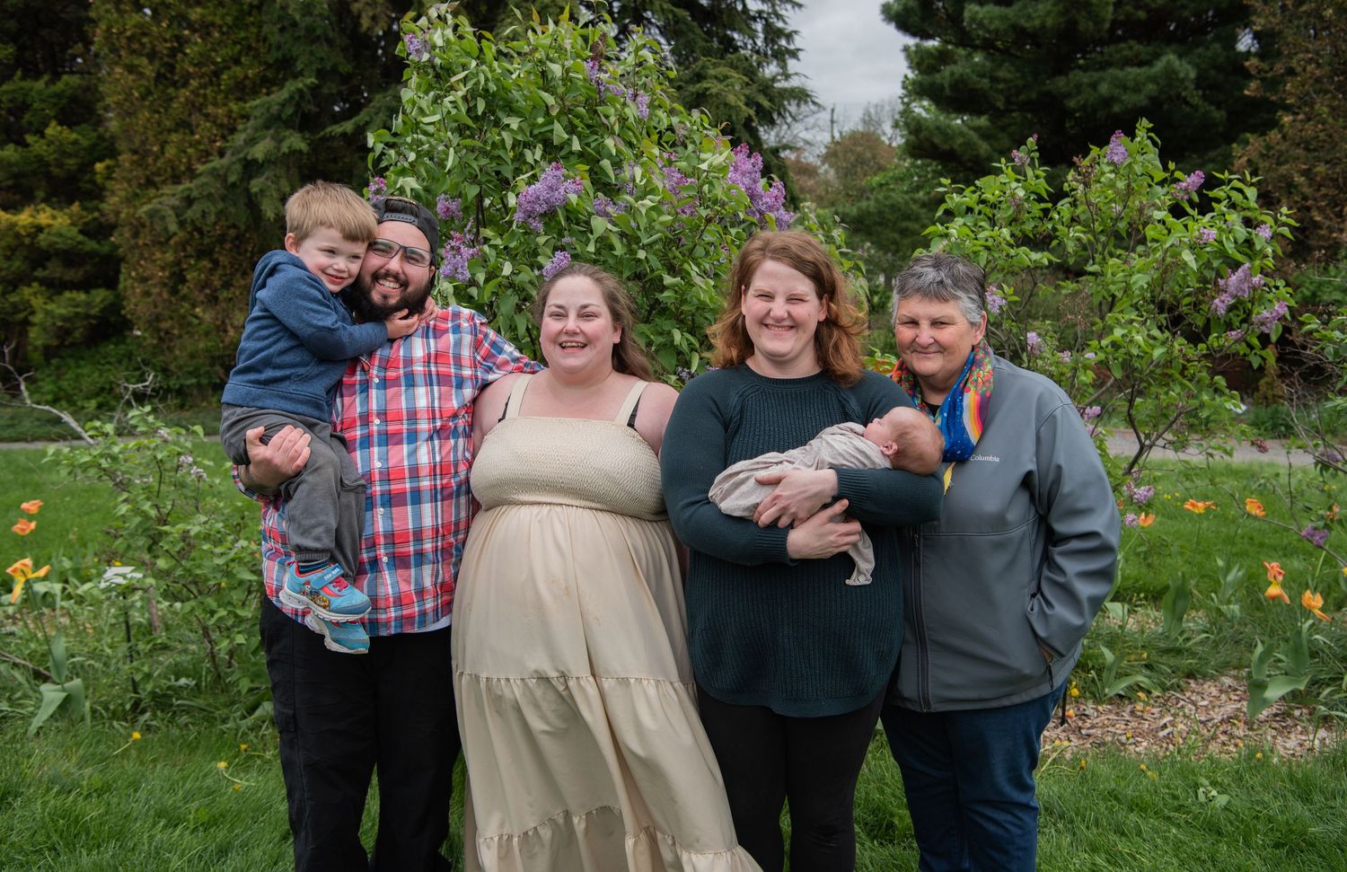 A multi-generational family group poses together in a garden with blooming flowers and greenery in the background.