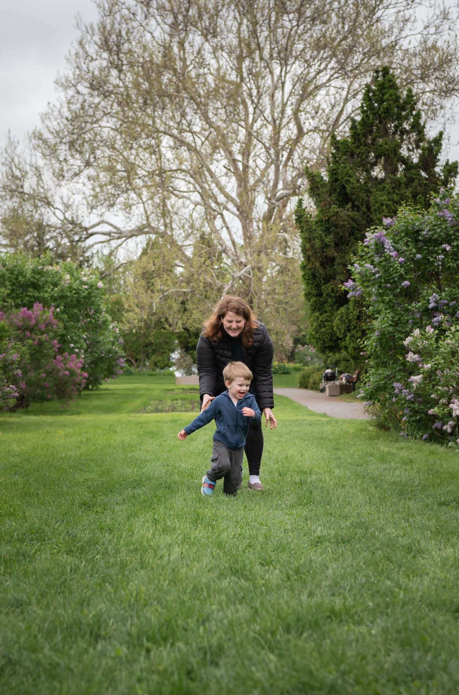 Two people enjoying a playful moment together while running on a grassy path lined with blooming lilacs and tall trees.