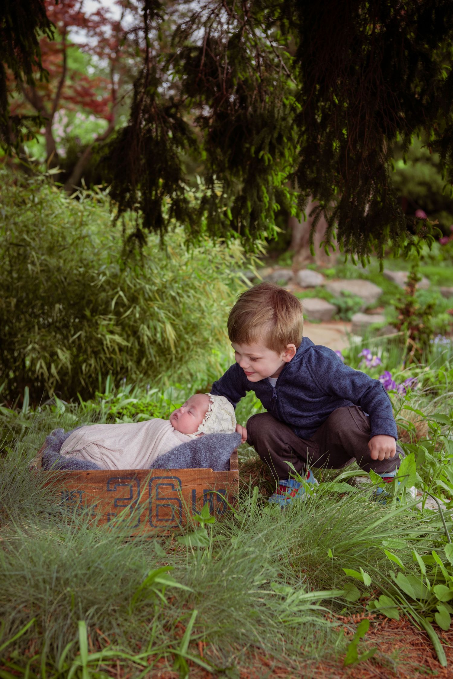 A young child crouches down to look at a baby resting in a woven basket outdoors among greenery and flowers.