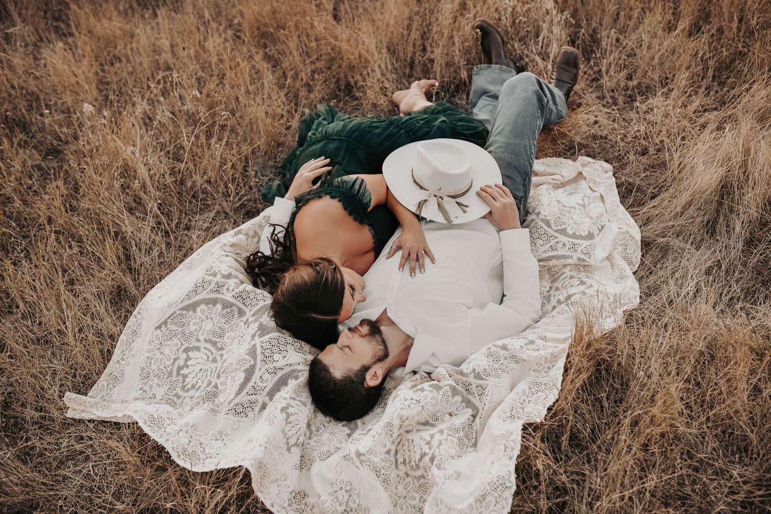a couple snuggles on a lace blanket in a field of golden grass during sunset for their engagement photos at murrell park