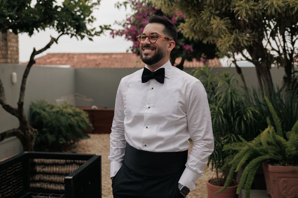A happy person wearing a crisp white dress shirt, black bow tie and black pants stands smiling in an outdoor garden setting.