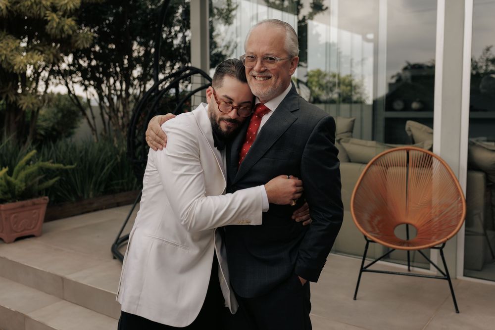 A heartfelt embrace between two people in formal attire, one in a white jacket and one in a black suit with red tie.