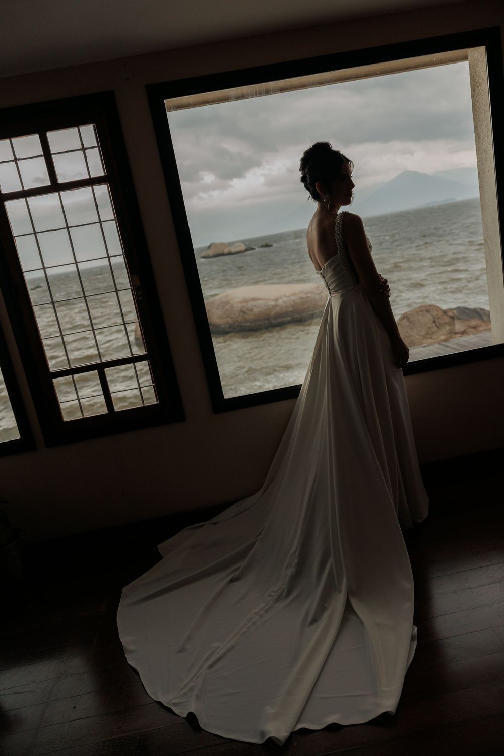 A person in a flowing white wedding dress stands silhouetted in front of a panoramic ocean view window.