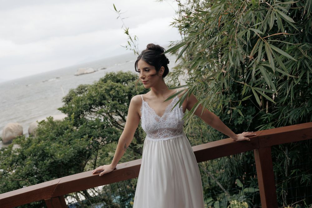 A bride in a flowing white v-neck dress stands on a balcony overlooking a scenic coastal landscape with greenery.