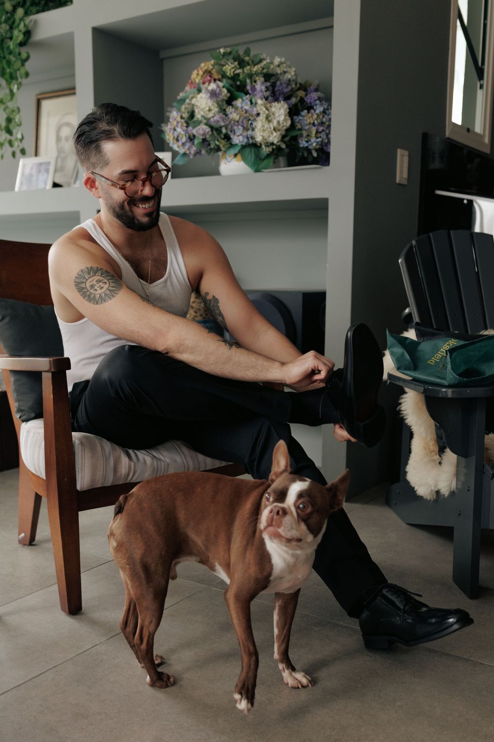 Brown and white Boston Terrier stands next to person sitting in wooden chair in modern living room with floral decor.