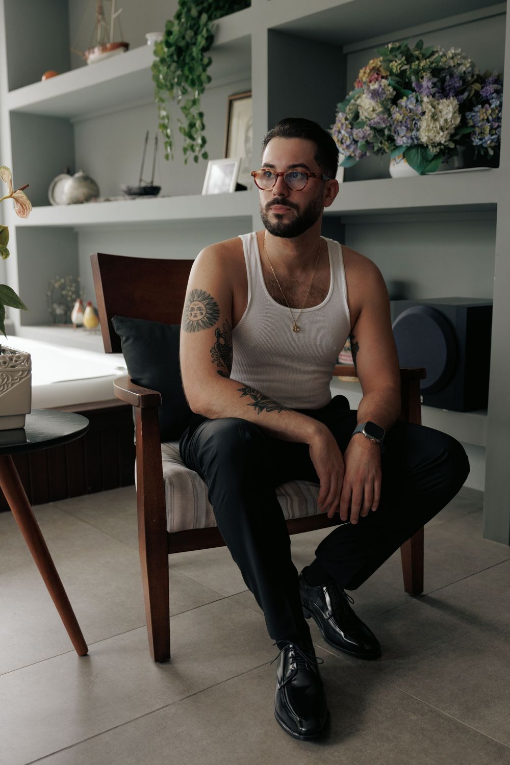 A person wearing a tank top and tattoos sits on a wooden chair against white built-in shelves with plants.