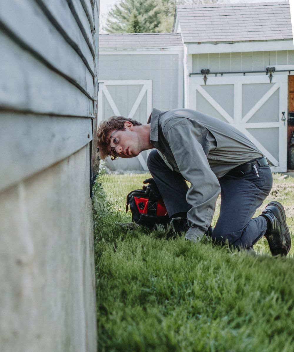A Modern Pest Control Technician kneeling in grass inspecting foundation or siding of house next to white barn doors in backyard.