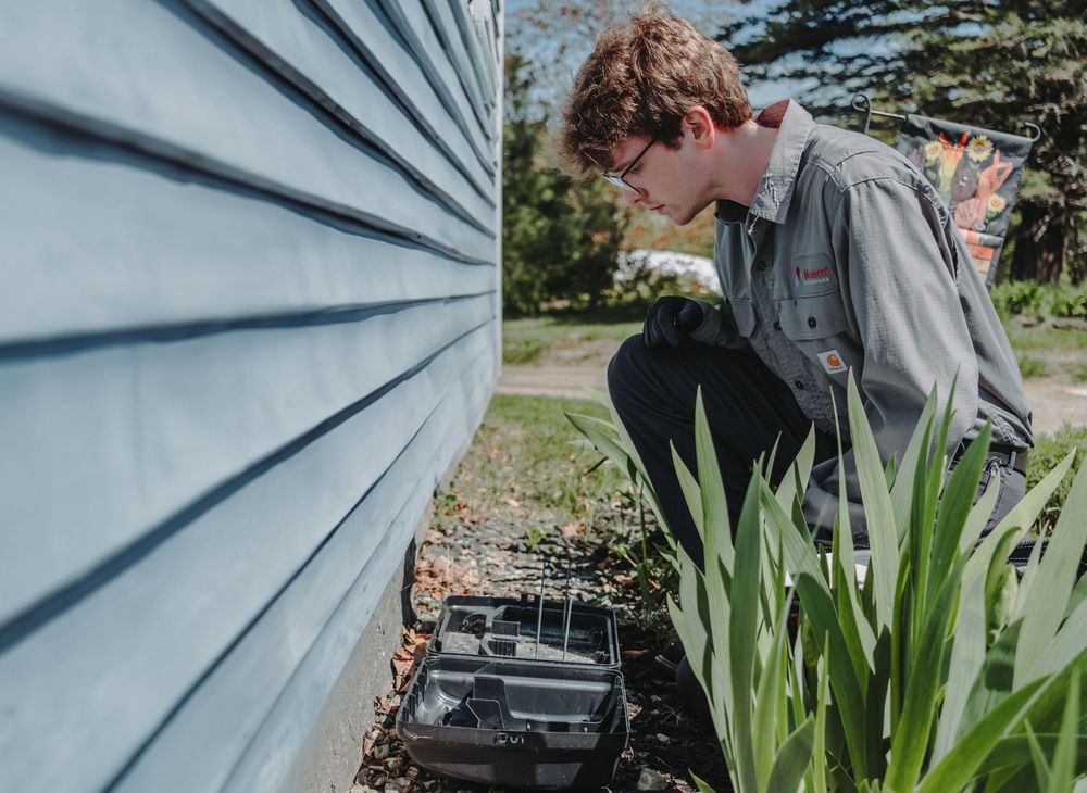 A Modern Pest Control Tech in a gray jacket bends down to inspect a rodent capture box along the side of a light blue house.