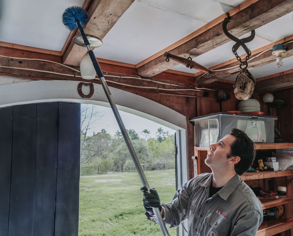 A Modern Pest Control Tech uses a professional duster with extension pole to clean cobwebs from the ceiling of a rustic wooden interior.
