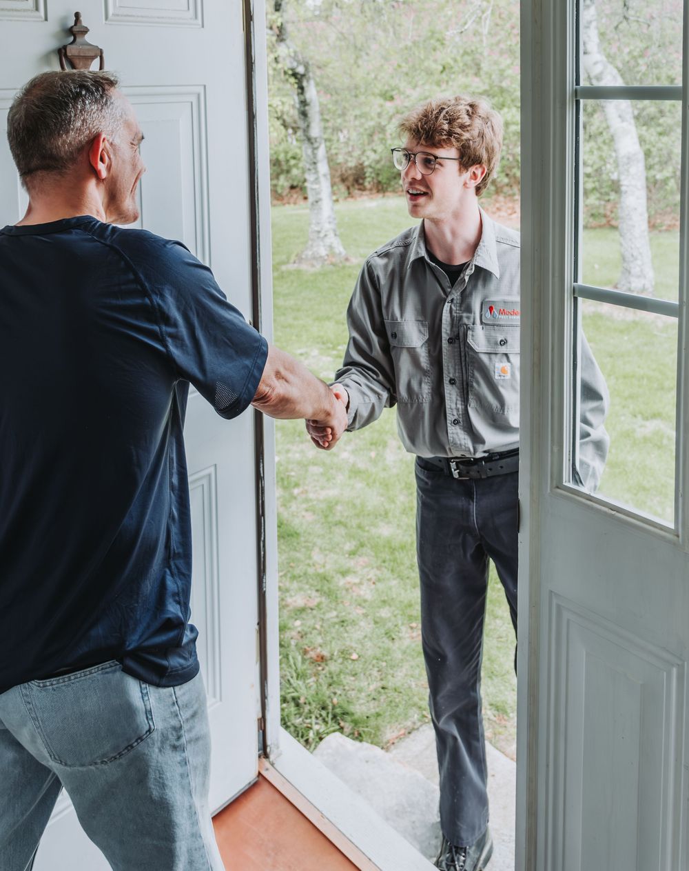 Two people shake hands at an open doorway with natural light and greenery visible. A Modern Pest Control Tech greets the homeowner.