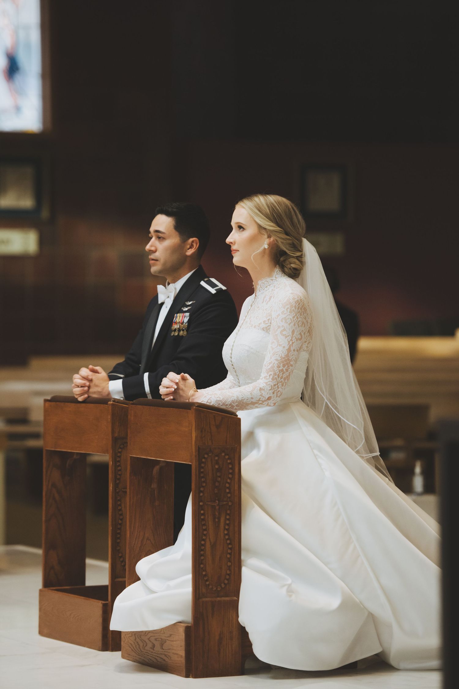 A couple in wedding attire kneels at wooden prayer benches during their church ceremony.