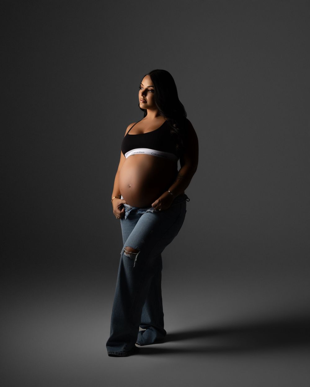 Silhouetted pregnant figure stands in jeans and crop top against dramatic dark backdrop in artistic maternity portrait.