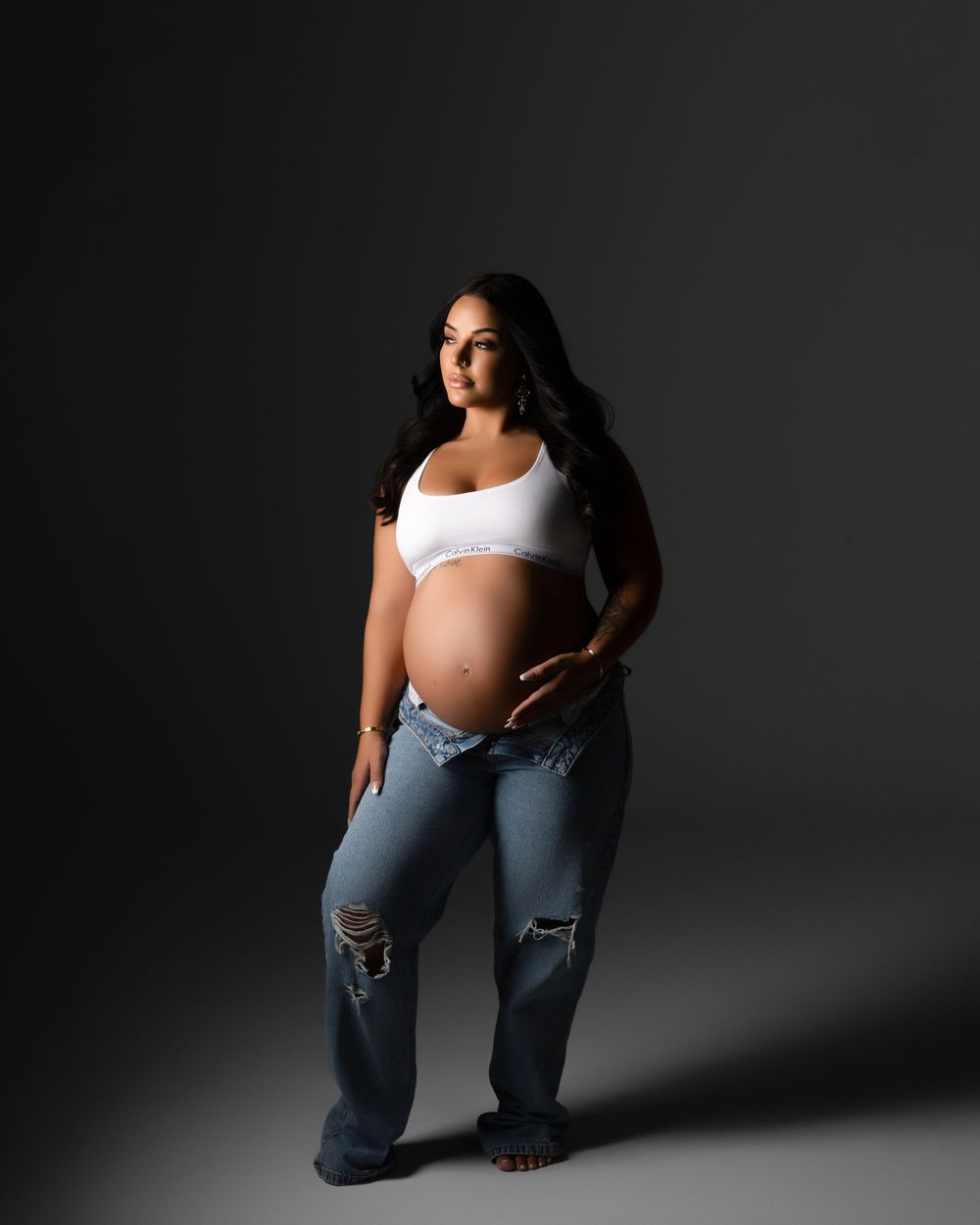 A woman in a white crop top and distressed baggy jeans poses against a dark studio backdrop in dramatic lighting.
