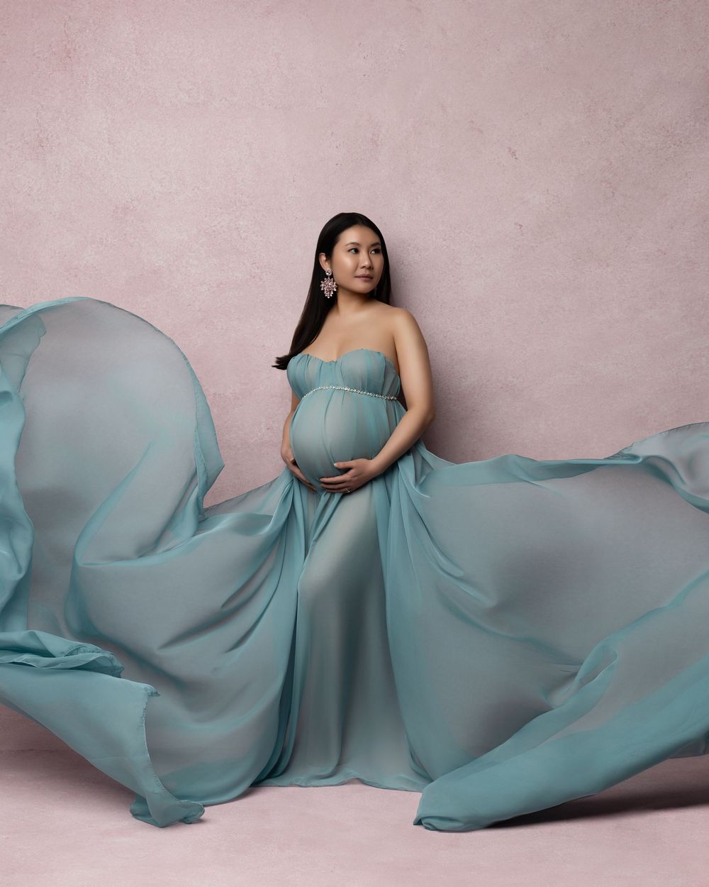 A flowing maternity photo series in light turquoise fabric showing a figure reclining against a dusty pink backdrop.