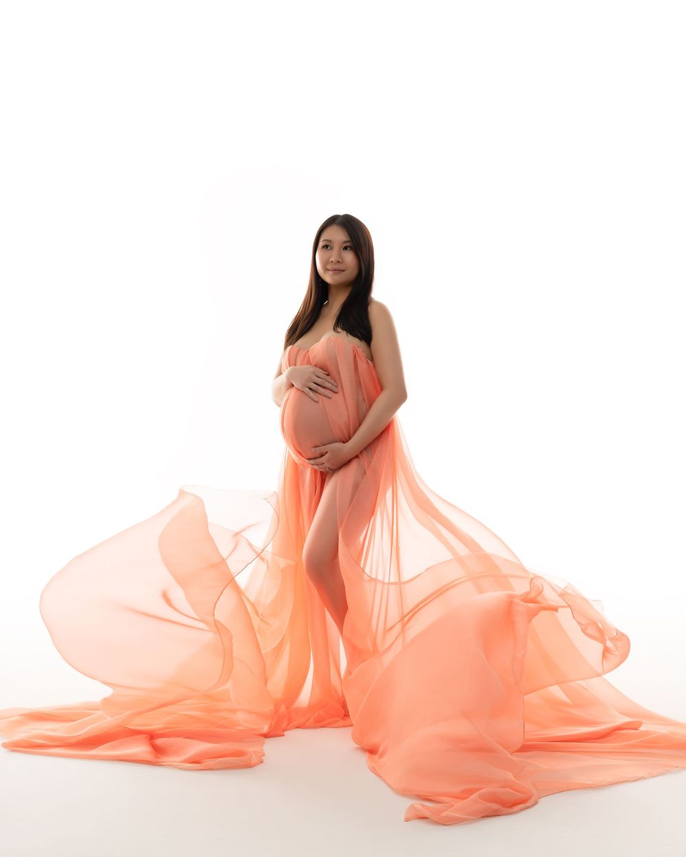 Expectant mother in peach-colored flowing maternity dress stands elegantly against white backdrop during photo session.