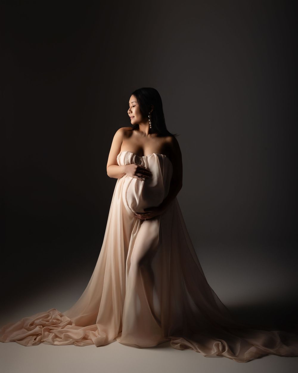 Dramatic maternity portrait in flowing blush gown against dark background with artistic lighting and elegant pose.