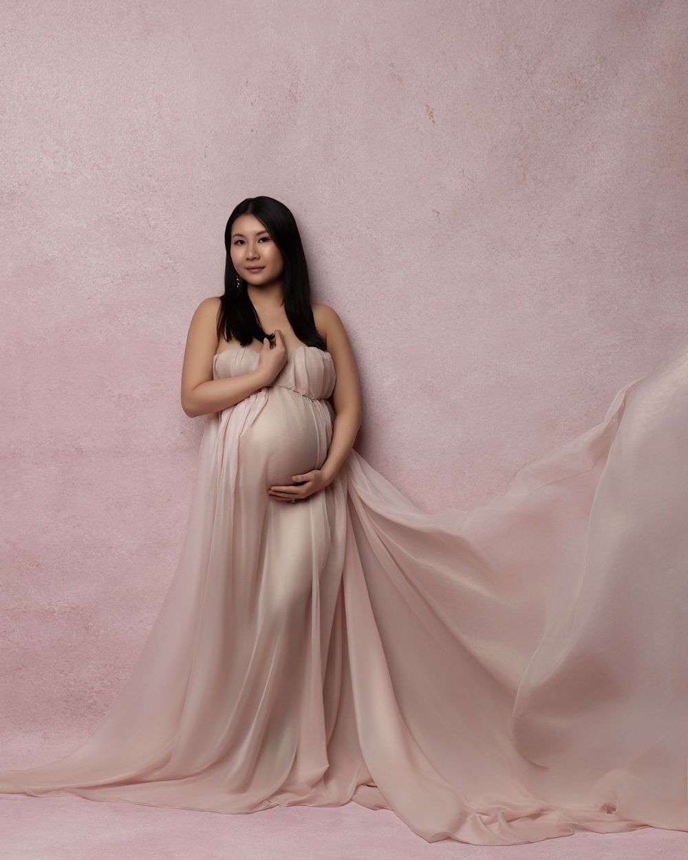 Expecting parent poses in flowing blush pink gown against textured pink backdrop during maternity photoshoot.