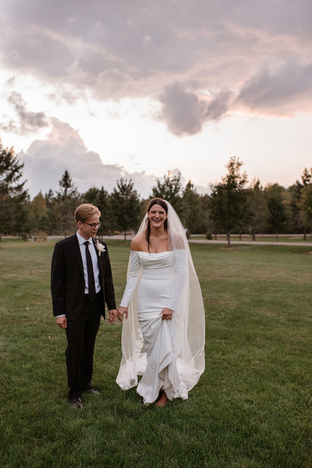 A romantic sunset wedding photo of newlyweds walking together on a grassy lawn with evergreen trees in the background.