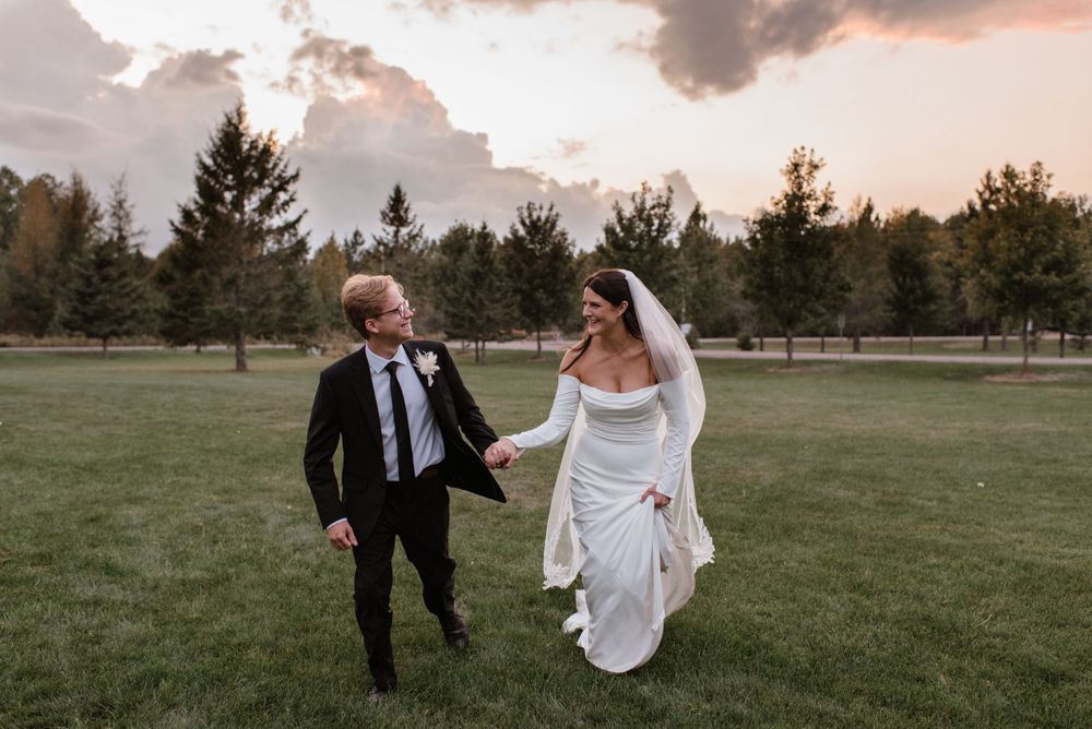 A couple in wedding attire runs joyfully across a grassy field at sunset with evergreen trees in the background.