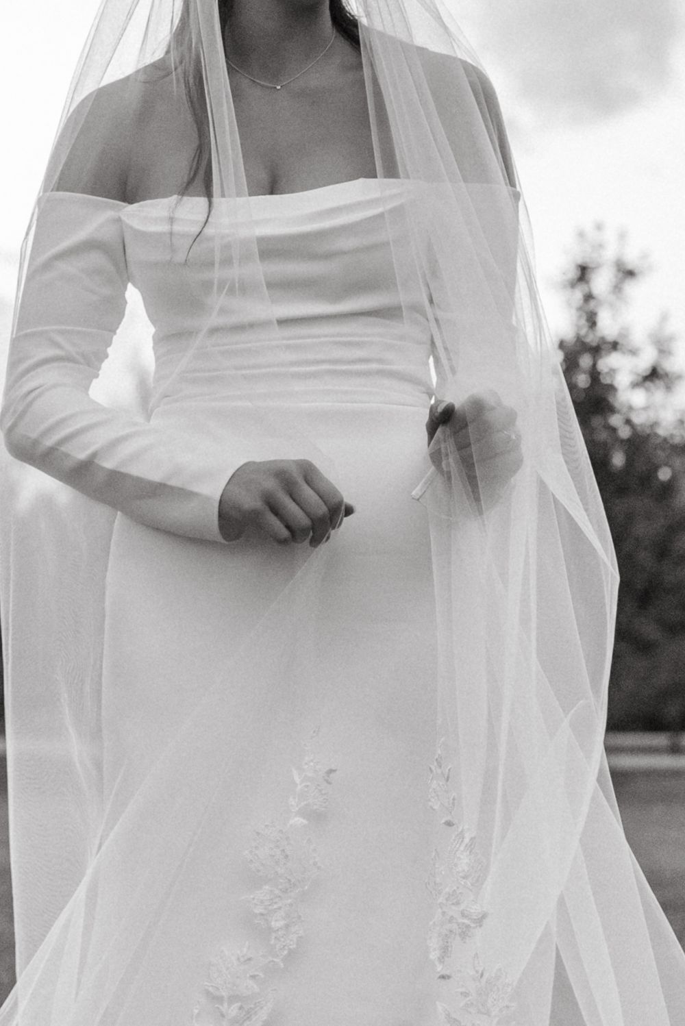 Black and white photo of a bride in a strapless wedding gown with long sleeves and flowing veil in an outdoor setting.