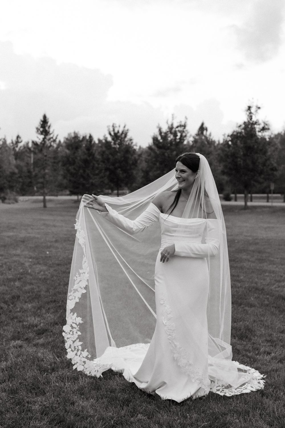 A black and white bridal portrait showing an elegant wedding dress and long flowing lace veil in a garden setting.