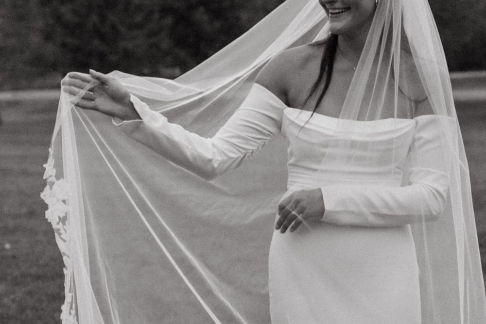 A bride in a long-sleeved white wedding dress gracefully adjusts her flowing veil outdoors in black and white photography.