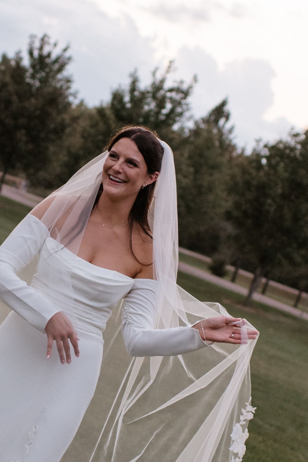 A bride in a white off-shoulder wedding dress poses outdoors while holding her flowing veil in a park setting.