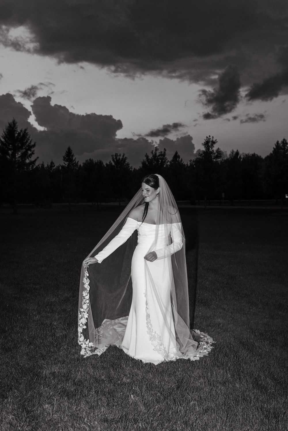 Black and white photo of a bride in an elegant dress with a long veil against a dramatic cloudy sunset.